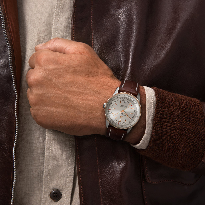 Juwelier-Morawitz-Blog-Breitling-Navitimer-3 Eine Person trägt eine braune Lederjacke und ein helles Hemd und trägt an ihrem linken Handgelenk eine elegante Analoguhr. Die Uhr vom Düsseldorfer Juwelier Morawitz hat ein braunes Lederarmband und ein silbernes Zifferblatt mit aufwendigen Details und strahlt einen klassischen und stilvollen Look aus.