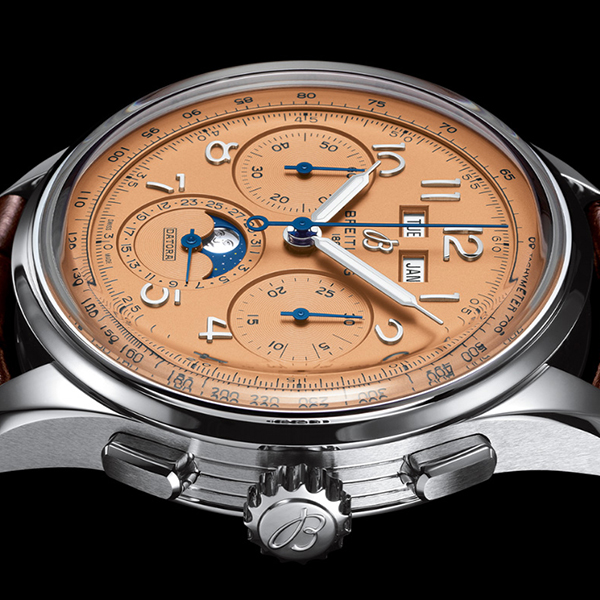 Nahaufnahme einer Luxusarmbanduhr mit detailliertem Zifferblatt. Das Zifferblatt, erhältlich bei Juwelier Morawitz in Düsseldorf, ist lachsfarben und verfügt über mehrere Hilfszifferblätter, eine Mondphasenanzeige sowie Tag-/Datumsfenster. Das Uhrengehäuse ist silberfarben mit einer geriffelten Krone und die Zeiger und Indizes sind in kontrastierendem Blau und Weiß gehalten.