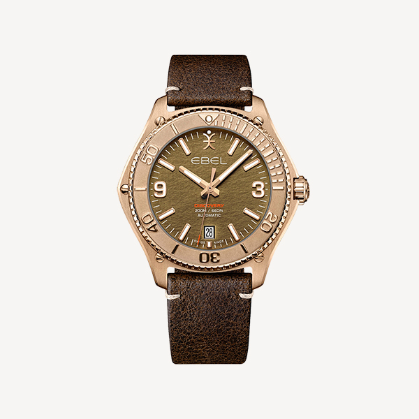 Eine Nahaufnahme einer Ebel-Armbanduhr von Juwelier Morawitz in Düsseldorf mit einem runden, bronzefarbenen Zifferblatt mit leuchtenden Markierungen und Zeigern sowie einer Datumsanzeige bei 6 Uhr. Die Uhr verfügt über ein braunes Lederarmband und eine bronzefarbene Lünette mit eingravierten Minutenmarkierungen auf einem schlichten weißen Hintergrund.