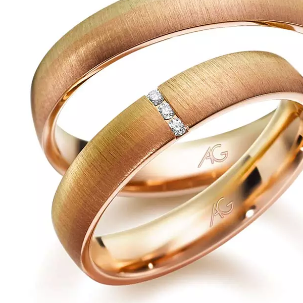 Juwelier-Morawitz-Box-Gerstner-Ansicht Abgebildet sind zwei Eheringe aus Roségold mit gebürsteter Oberfläche. Der größere Ring ist schlicht, während der kleinere Ring mit drei kleinen, in einer Reihe angeordneten Diamanten besetzt ist. Beide Ringe wurden von Juwelier Morawitz in Düsseldorf gefertigt und haben auf der Innenseite eine „AG“-Gravur.