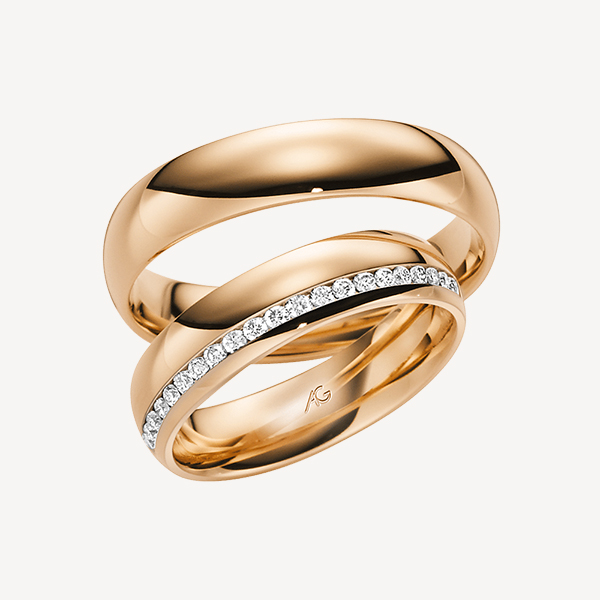 Vor einem schlichten Hintergrund ist ein Paar Goldringe von Juwelier Morawitz aus Düsseldorf zu sehen. Ein Ring ist ein schlichter Goldring, während der andere ähnlich gestaltet ist und eine Reihe kleiner Diamanten aufweist, die entlang seines Umfangs eingelassen sind. Die Ringe sind so angeordnet, dass einer in dem anderen liegt.