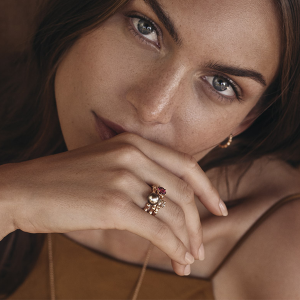 Juwelier-Morawitz-Box-Joerg-Heinz-Ansicht Nahaufnahme des Gesichts einer Frau mit hellen Augen und braunem Haar, die ihr Kinn auf ihre Hand stützt, die mit einem goldenen Ring von Juwelier Morawitz in Düsseldorf geschmückt ist. Sie hat einen ruhigen Gesichtsausdruck und trägt ein ärmelloses braunes Oberteil, aus dem eine minimalistische goldene Halskette hervorschaut.