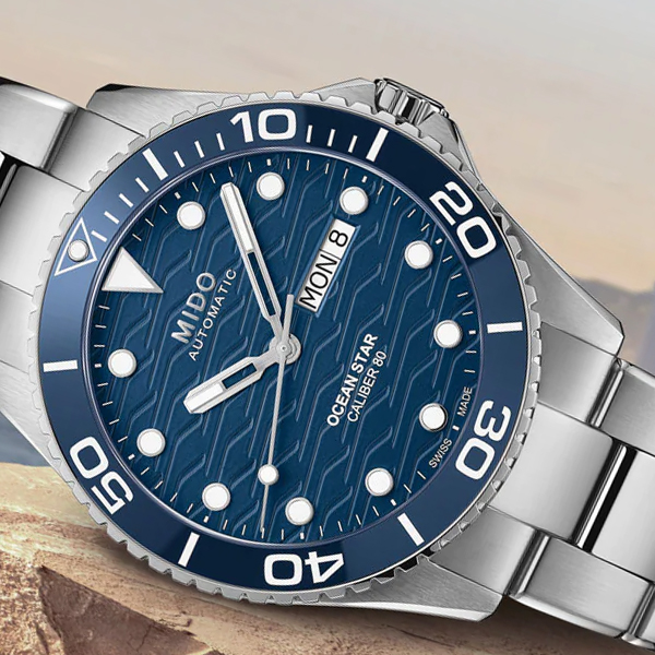 Eine elegante silberne Armbanduhr mit blauem Zifferblatt mit Wellenmuster, weißen Akzenten und fettgedruckten Ziffern, die alle zehn Minuten markieren. Oben steht der Markenname „MIDO“, unten die Worte „OCEAN STAR CALIBER 80“. Diese Uhr ist exklusiv bei Juwelier Morawitz in Düsseldorf erhältlich und zeigt auch Tag und Datum an.