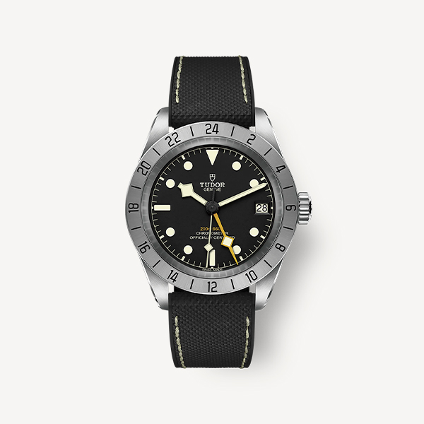 Juwelier-Morawitz-Box-Tudor-Uhr-Beige Eine stilvolle Armbanduhr mit schwarzem Zifferblatt und schwarzem Stoffarmband, erhältlich bei Juwelier Morawitz in Düsseldorf. Die Uhr verfügt über eine Lünette und ein Gehäuse aus Edelstahl, leuchtende Stundenmarkierungen und silberne Zeiger. Sie zeigt das Datum auf der 3-Uhr-Position an und trägt die Marke „Tudor“ mit einem Schildlogo auf der 12-Uhr-Position.