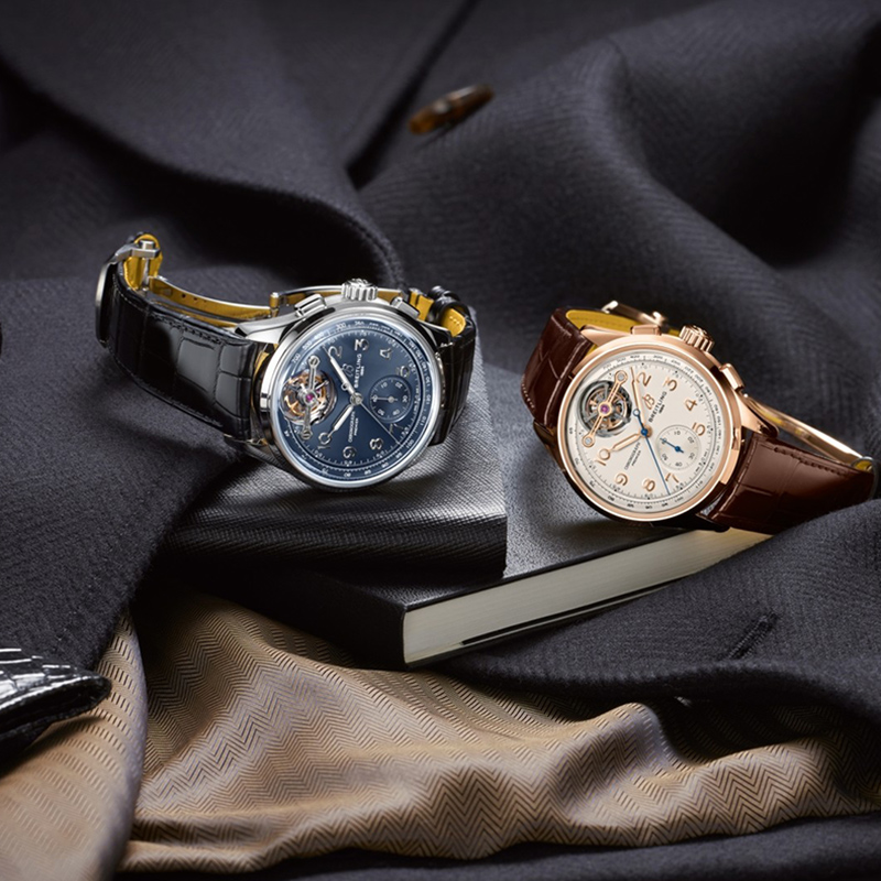 Juwelier-Morawitz-Breitling-Uhren-Vorschau-2022 Zwei Luxusarmbanduhren von Juwelier Morawitz aus Düsseldorf werden auf einem dunkel strukturierten Mantel und beigem Stoff präsentiert. Eine Uhr hat ein schwarzes Lederarmband und ein blaues Zifferblatt, während die andere ein braunes Lederarmband und ein weißes Zifferblatt hat. Beide haben komplizierte Designs und liegen auf einem geschlossenen Buch.