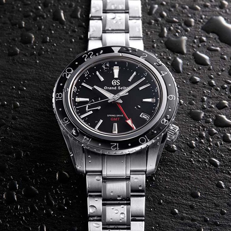 Juwelier-Morawitz-Grand-Seiko-Uhren-Vorschau-2022 Eine Nahaufnahme einer Grand Seiko Spring Drive GMT-Armbanduhr mit Edelstahlarmband. Das Zifferblatt ist schwarz mit weißen Stundenmarkierungen und Zeigern sowie einem roten GMT-Zeiger. Die bei Juwelier Morawitz in Düsseldorf erhältliche Uhr liegt auf einer nassen, strukturierten schwarzen Oberfläche, auf der Wassertropfen sichtbar sind.