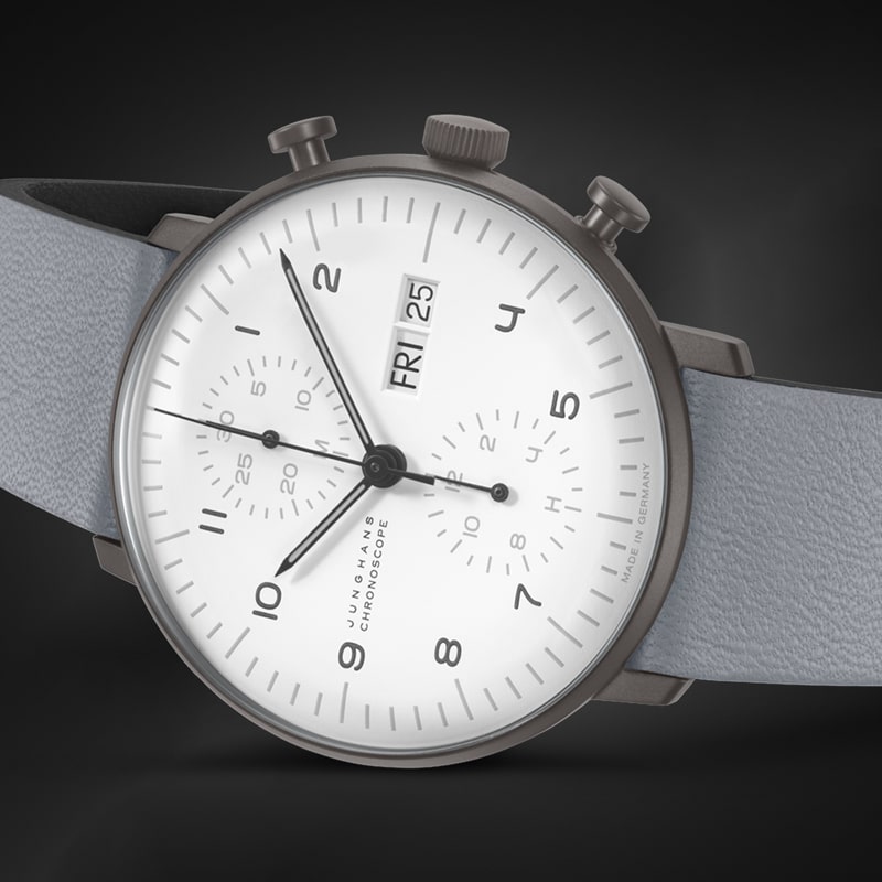 Juwelier-Morawitz-Junghans-Uhren-Vorschau-2 Eine elegante, moderne Armbanduhr von Juwelier Morawitz mit weißem Zifferblatt mit schwarzen Ziffern und Zeigern. Die Uhr zeigt Tag und Datum an und verfügt über drei Hilfszifferblätter zum Anzeigen von Sekunden, Minuten und Stunden. Sie hat ein hellgraues Lederarmband und ein Metallgehäuse vor einem dunklen Düsseldorfer Hintergrund.