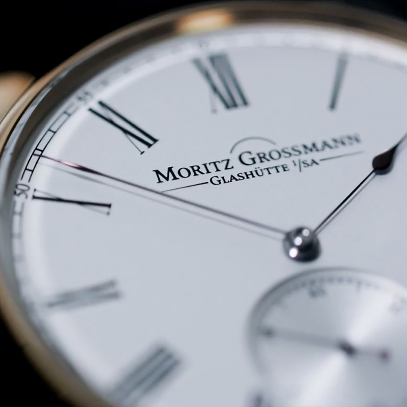 Juwelier-Morawitz-Moritz-Grossmann-Anzeige-2 Nahaufnahme einer Armbanduhr von Moritz Grossmann Glashütte von Juwelier Morawitz in Düsseldorf. Die Uhr verfügt über ein weißes Zifferblatt mit schwarzen römischen Stundenmarkierungen und schmalen Zeigern. Der Markenname ist in der Mitte deutlich zu erkennen, und ein Hilfszifferblatt für den Sekundenzeiger ist auf der 6-Uhr-Position sichtbar.