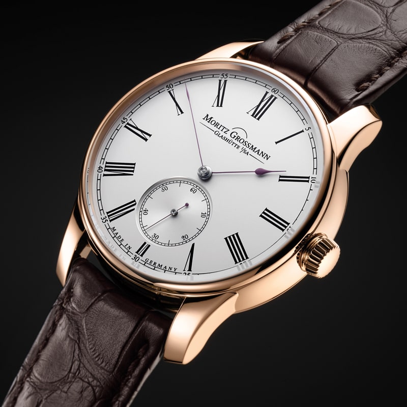 Juwelier-Morawitz-Moritz-Grossmann-Beitragsbild Eine Luxusarmbanduhr mit goldenem Gehäuse, weißem Zifferblatt und braunem Lederarmband. Das Zifferblatt verfügt über schwarze römische Ziffern, Minutenmarkierungen und ein kleines Sekundenzifferblatt über der 6-Uhr-Position. Der Markenname „Moritz Grossmann“ ist elegant unter der 12-Uhr-Position angebracht und ist bei Juwelier Morawitz in Düsseldorf zu finden.