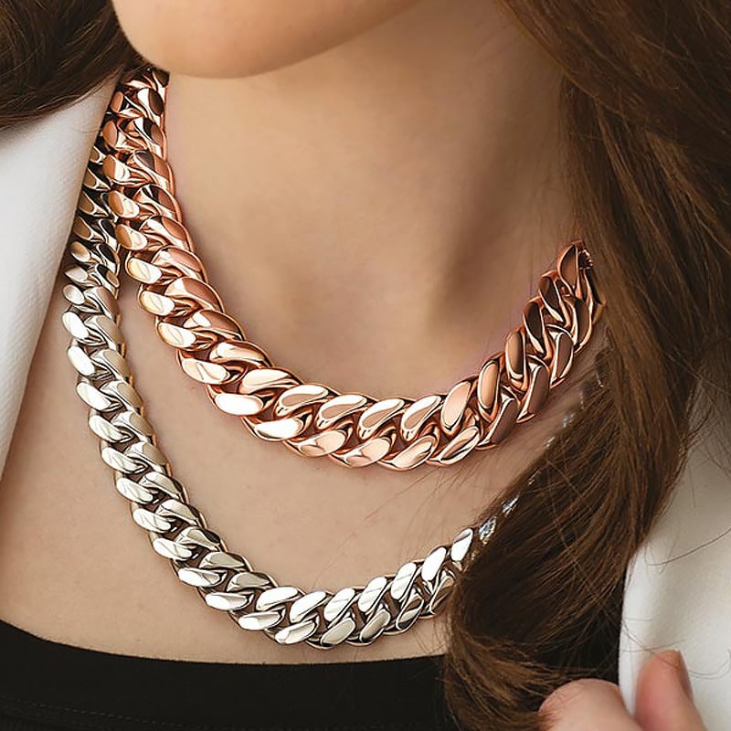 Juwelier-Morawitz-Schmuck-Emil-Kraus-Ansicht-2 Eine Nahaufnahme einer Person, die zwei klobige, metallische Kettenhalsketten von Juwelier Morawitz in Düsseldorf trägt. Eine Kette ist aus Roségold und die andere aus Silber, wobei die Roségoldkette über der Silberkette liegt. Die Person trägt ein schwarzes Oberteil und eine weiße Jacke.