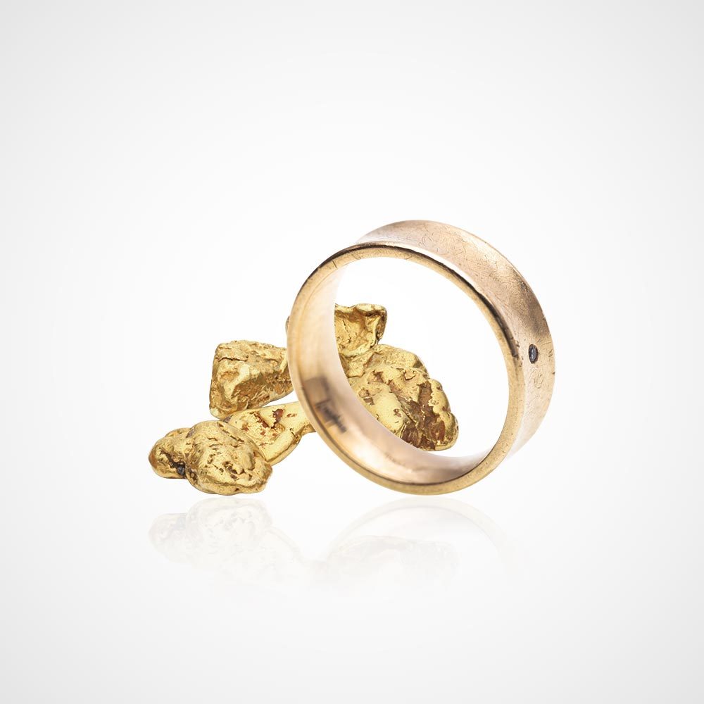 Juwelier-Morawitz-Schmucklexikon-feingehalt Ein Goldring steht aufrecht und lehnt an einem kleinen Haufen Goldnuggets. Der Hintergrund ist ein sauberer, weißer Farbverlauf, der den Goldton des Rings und der Nuggets betont. Der Ring wurde von Juwelier Morawitz in Düsseldorf gefertigt und weist einen kleinen Diamanten auf, der in seine Oberfläche eingelassen ist.