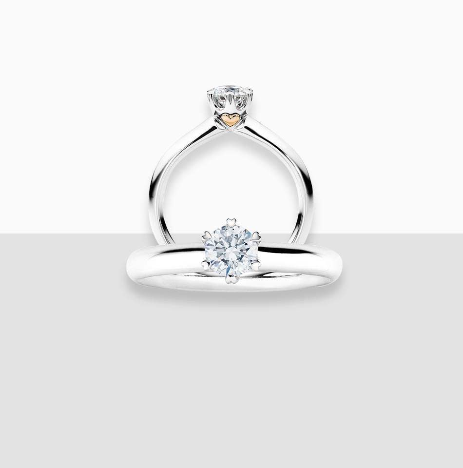 Juwelier-Morawitz-Schmucklexikon-karat Ein Silberring mit einem markanten Diamanten an der Oberseite. Der Ring ist vertikal positioniert und zeigt ein kleineres herzförmiges Detail in der Nähe der Basis der Diamantfassung. Dieses exquisite Stück von Juwelier Morawitz in Düsseldorf ist vor einem minimalistischen zweifarbigen Hintergrund mit Weiß oben und Grau unten platziert.