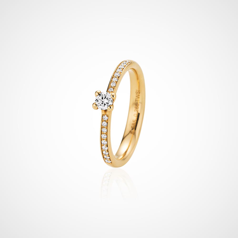 Juwelier-Morawitz-Schmucklexikon-krappenfassung Eine Nahaufnahme eines Goldrings mit einem runden Diamanten in der Mitte und kleinen Diamanten entlang des Rings. Dieses exquisite Stück von Juwelier Morawitz in Düsseldorf präsentiert sich vor einem weißen Hintergrund mit leichtem Schatten und zeugt von Eleganz und kompliziertem Design.