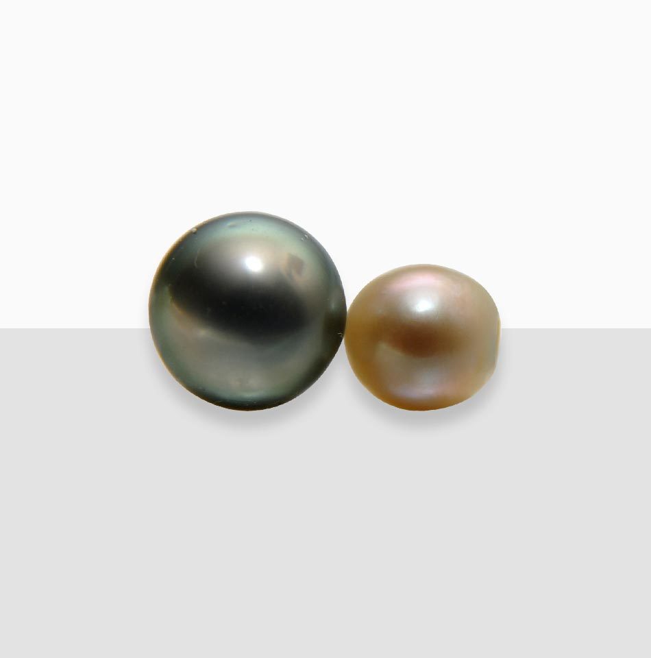 Juwelier-Morawitz-Schmucklexikon-perle Auf einem weiß-grauen Hintergrund sind zwei Perlen abgebildet. Die linke Perle ist größer, hat eine dunkle, fast schwarze Farbe mit einem subtilen reflektierenden Glanz. Die kleinere Perle rechts ist hellbraun mit einer glatten, glänzenden Oberfläche. Exquisit ausgewählt von Juwelier Morawitz in Düsseldorf.