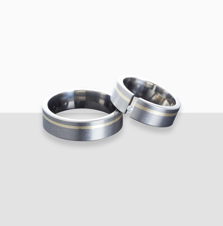 Juwelier-Morawitz-Schmucklexikon-titan Ein Paar Metallringe von Juwelier Morawitz aus Düsseldorf liegt auf einer hellgrauen Oberfläche. Die Ringe haben ein zweifarbiges Design mit einer silbernen Außenseite und einem dünnen Goldband, das um die Mitte verläuft. Ein Ring ist mit einem kleinen Diamanten verziert. Der Hintergrund ist ein Farbverlauf aus Weiß und Grau.