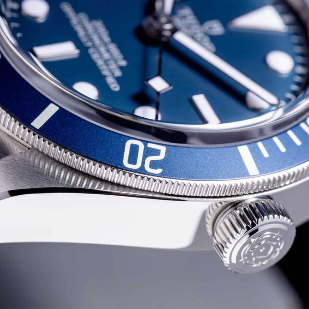 Juwelier-Morawitz-Uhrenlexikon-krone Nahaufnahme einer Luxusarmbanduhr mit blauem Zifferblatt und Lünette, erhältlich bei Juwelier Morawitz in Düsseldorf. Die Uhr verfügt über leuchtende Stundenmarkierungen und Zeiger sowie eine seitliche Krone mit eingraviertem Logo. Die Lünette ist mit Ziffern versehen und ein Teil des Gehäuses ist sichtbar und zeigt seine polierte Oberfläche.