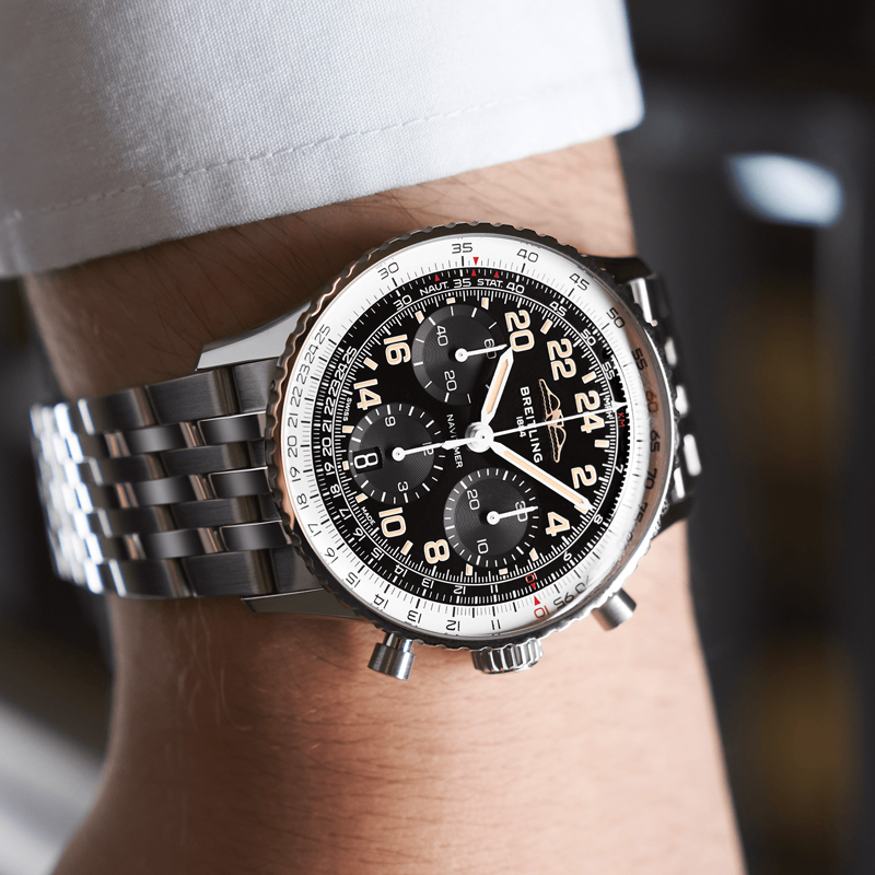 Juwelier-Morawitz-Breitling-Uhren-Vorschau-2022-12 Eine Nahaufnahme eines Handgelenks, an dem eine luxuriöse Breitling Chronomat-Uhr von Juwelier Morawitz in Düsseldorf getragen wird. Die Uhr hat ein schwarzes Zifferblatt mit mehreren Hilfszifferblättern, eine 24-Stunden-Zeitanzeige und ein Edelstahlarmband. Die Lünette ist silberfarben mit eingravierten Ziffern vor einem wunderschön verschwommenen Hintergrund.