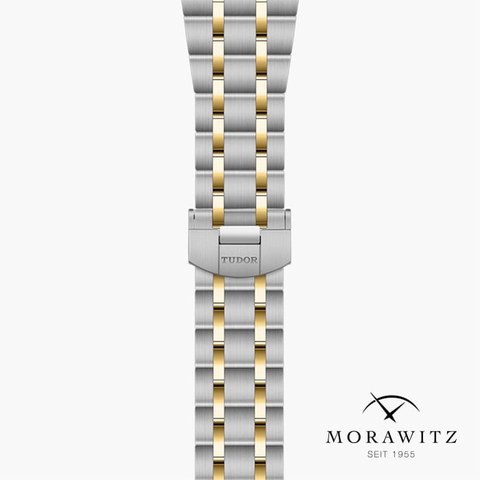 Eine Nahaufnahme eines zweifarbigen Metallarmbands mit silbernen und goldenen Gliedern. In die Schließe ist das Wort „Tudor“ eingraviert. In der unteren rechten Ecke sind das Logo und der Name „Morawitz“ mit dem Text „Seit 1955“ zu sehen.
