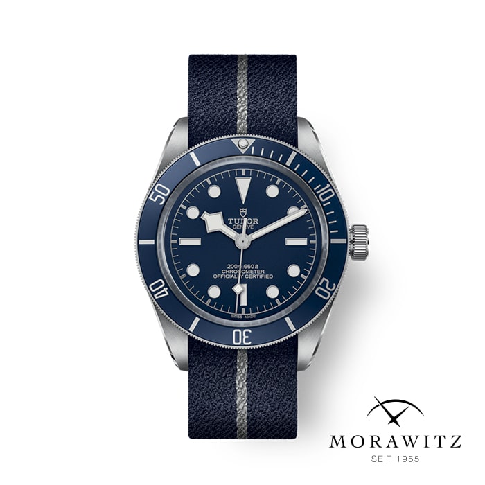 Eine stilvolle Armbanduhr mit tiefblauem Zifferblatt und Lünette, leuchtenden Stundenmarkierungen und Zeigern sowie einem gestreiften blauen Stoffarmband. Die Marke „Tudor“ ist auf dem Zifferblatt zu sehen. Das Bild enthält in der unteren rechten Ecke das Logo „MORAWITZ SEIT 1955“.
