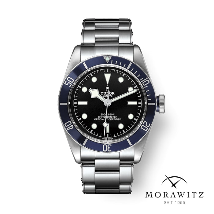 Eine Tudor-Uhr mit schwarzem Zifferblatt, silberfarbenem Edelstahlarmband und schwarzer Lünette mit Minutenmarkierungen. Die Stundenmarkierungen sind Punkte, mit einem Dreieck auf der 12-Uhr-Position. Die Uhr ist auf einem weißen Hintergrund mit dem Morawitz-Logo und dem Text „SEIT 1955“ in der unteren rechten Ecke angebracht.