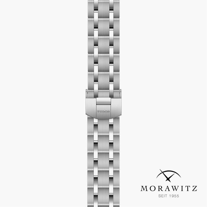Nahaufnahme eines silbernen Uhrenarmbands aus Metall mit ineinandergreifenden Segmenten. In die mittlere Schließe ist der Markenname „Tudor“ eingraviert. In der unteren rechten Ecke sind der Markenname „Morawitz“ und der Satz „Seit 1955“ in schwarzer Schrift zu sehen.