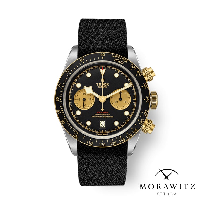 Eine luxuriöse Tudor-Armbanduhr mit schwarz-goldenem Zifferblatt, schwarzem Stoffarmband, goldenen Chronographenknöpfen und Datumsanzeige bei 6 Uhr. Das Morawitz-Logo und der Text „Seit 1955“ sind in der unteren rechten Ecke sichtbar.