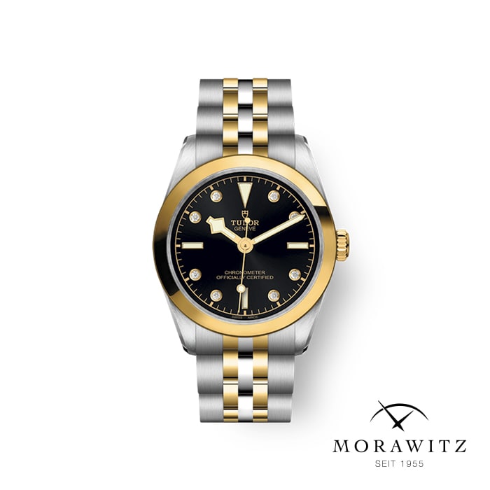 Eine Tudor-Armbanduhr mit schwarzem Zifferblatt, goldenen Akzenten und einem zweifarbigen Armband aus Edelstahl in Gold und Silber ist vor einem schlichten weißen Hintergrund zu sehen. Das Morawitz-Logo und der Text „seit 1955“ sind in der unteren rechten Ecke zu sehen.