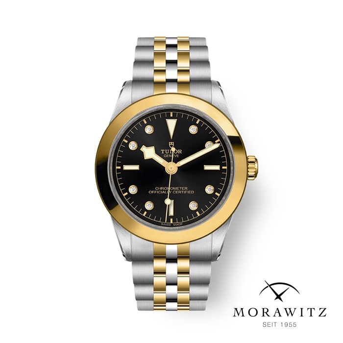 Eine Luxusarmbanduhr mit schwarzem Zifferblatt, goldenen Akzenten, Diamant-Stundenmarkierungen und einem zweifarbigen Metallarmband. Der Markenname „Tudor“ ist auf dem Zifferblatt zu sehen. Die Uhr ist auf einem weißen Hintergrund platziert und hat unten rechts das Logo „Morawitz Seit 1955“.