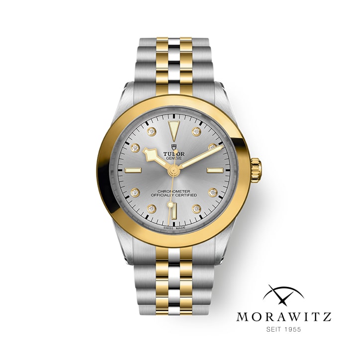 Eine Armbanduhr mit einem silber- und goldfarbenen Metallarmband. Die Uhr verfügt über ein silbernes Zifferblatt mit goldenen Stundenmarkierungen und Zeigern. Der Markenname „Tudor“ ist unterhalb der 12-Uhr-Position sichtbar. Das Bild enthält unten rechts ein „Morawitz“-Logo und den Text „Seit 1955“.