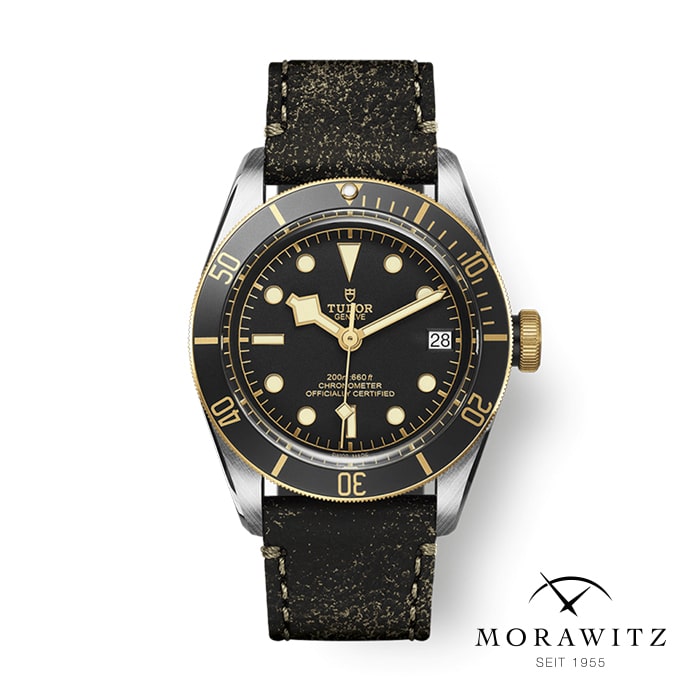 Eine Tudor-Armbanduhr mit schwarzer und goldener Lünette, schwarzem Zifferblatt und goldenen Akzenten wird ausgestellt. Die Uhr verfügt über ein Datumsfenster, Leuchtmarkierungen und ein schwarzes strukturiertes Armband. Das Morawitz-Logo mit „SEIT 1955“ befindet sich in der unteren rechten Ecke.