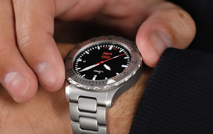 Eine Person trägt eine silberne Armbanduhr mit schwarzem Zifferblatt und roten Akzenten. Die Uhr hat ein Metallarmband und die Person stellt die Zeit mit einem Zeiger ein. Sie trägt ein dunkles Langarmhemd.