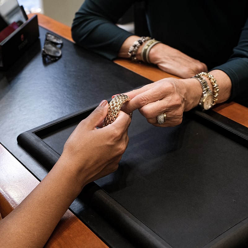 Juwelier-Morawitz-Uhren-Schmuck-Duesseldorf-Contentbild-1 Zwei Personen an einem Schmuckstand, eine probiert ein goldenes Armband mit Gittermuster an. Beide tragen Uhren und Armbänder. Auf dem Stand stehen eine Brille und ein schwarzes Tablett, eine Person zeigt auf das Armband.
