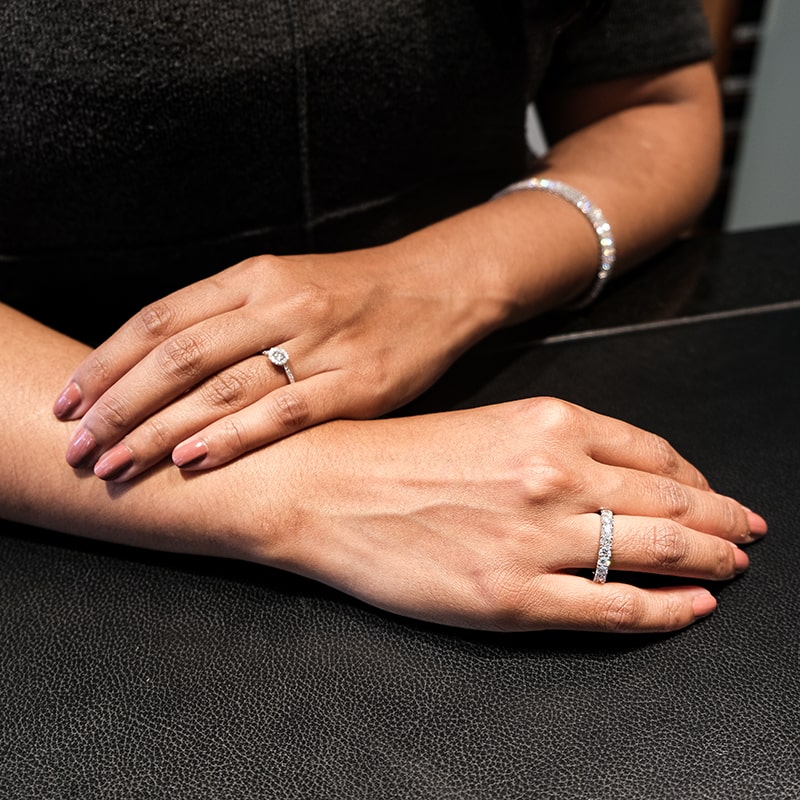 Juwelier-Morawitz-Uhren-Schmuck-Duesseldorf-Contentbild-3 Zwei Hände ruhen auf einer dunklen Oberfläche. Eine Hand trägt einen Diamantring und ein silbernes Armband, die andere einen Ehering mit Diamanten. Die Nägel sind rosa lackiert. Der Hintergrund ist verschwommen und dunkel.