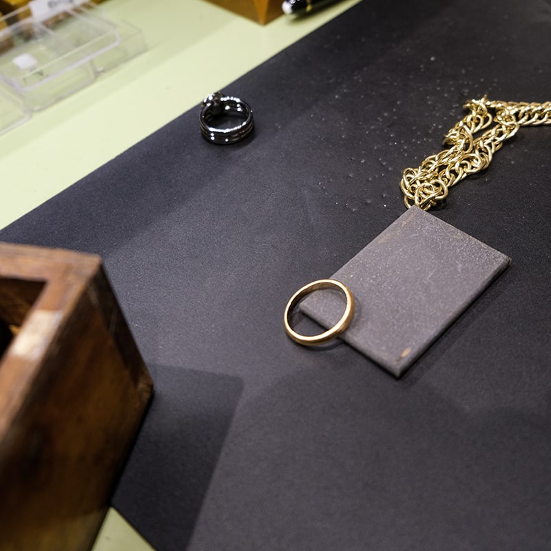 Juwelier-Morawitz-Uhren-Schmuck-Duesseldorf-Contentbild-6 Eine goldene Kette mit einem großen, flachen Anhänger und einem runden Ring liegt auf einer dunklen Oberfläche. In der Nähe sind eine Holzkiste und ein schwarzer Ring zu sehen. Im Hintergrund sind ein Stück Papier und ein durchsichtiger Behälter zu sehen.