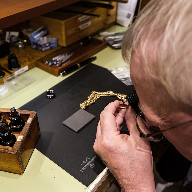 Juwelier-Morawitz-Uhren-Schmuck-Duesseldorf-Contentbild-7 Eine Person untersucht eine Goldkette mit einer Lupe. Am Arbeitsplatz sind Tintenfässer, ein Smartphone und verschiedene Werkzeuge zu sehen. Die Umgebung lässt auf eine sorgfältige Inspektion oder Reparatur in einer Werkstatt oder einem Arbeitsbereich schließen.