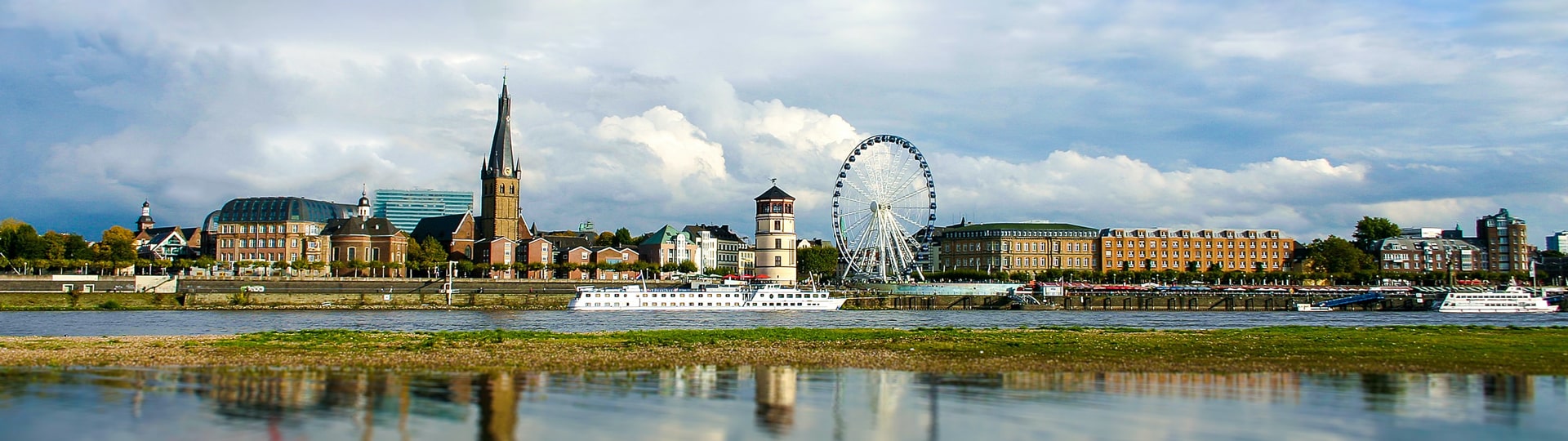 Eine malerische Aussicht auf die Skyline einer Stadt mit historischen Gebäuden, einer markanten Kirche mit einem hohen Turm, einem Riesenrad und einem Fluss im Vordergrund. Ein paar Boote segeln unter einem teilweise bewölkten Himmel auf dem Wasser.