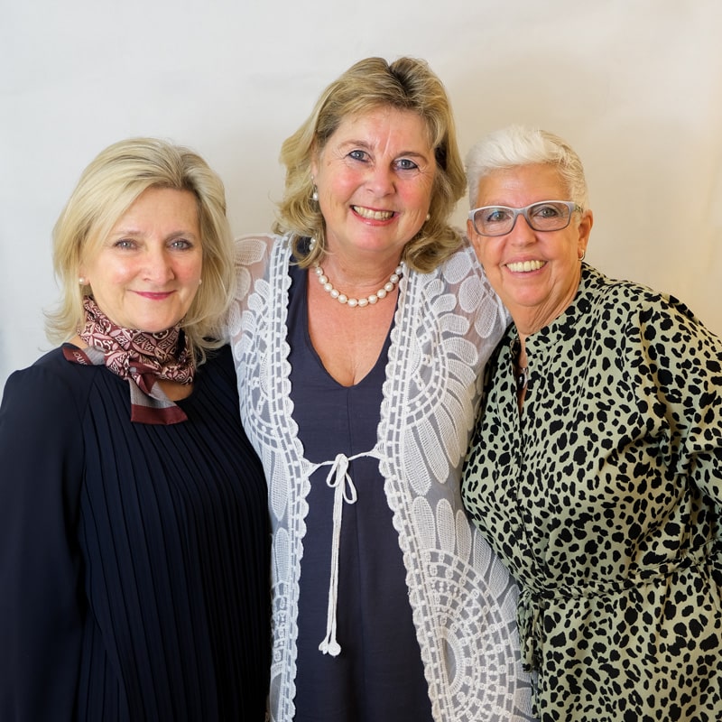 Ihre Ansprechpartner bei Juwelier Morawitz Drei Frauen stehen dicht beieinander und lächeln. Die Frau links trägt ein schwarzes Kleid mit Schal, die Frau in der Mitte ein helles Kleid mit Spitzendetails und die Frau rechts trägt eine Brille und ein Outfit mit Leopardenmuster. Der Hintergrund ist schlicht weiß.