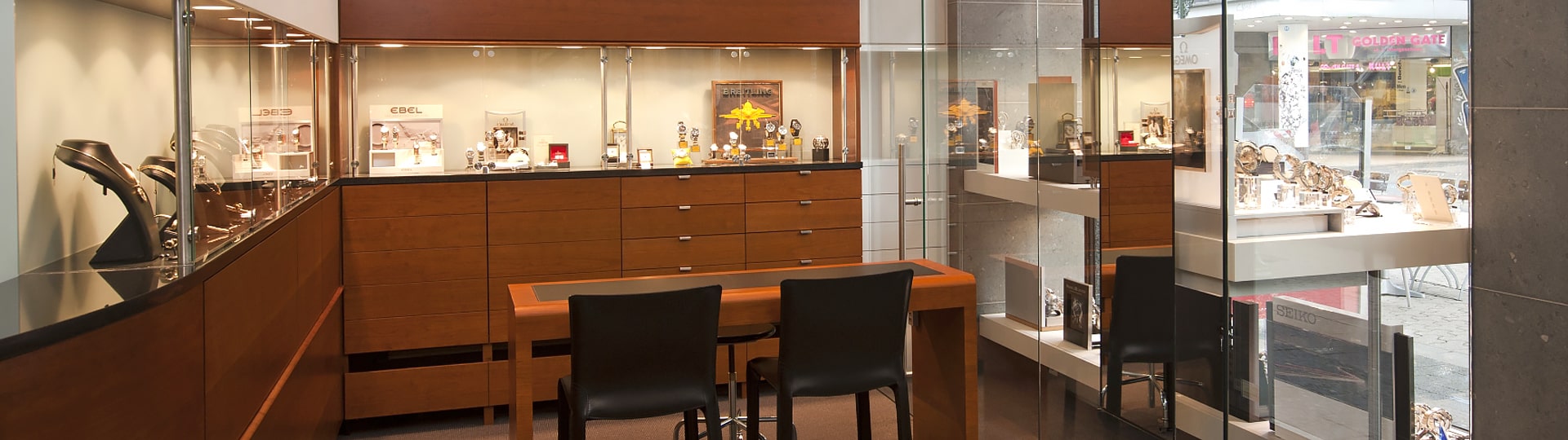 Innenansicht eines Uhrengeschäfts mit einem Holztisch und zwei Stühlen in der Mitte. Glasvitrinen präsentieren verschiedene Uhren im Raum. Große Fenster sorgen für Tageslicht und bieten einen Blick auf den Einkaufsbereich draußen.