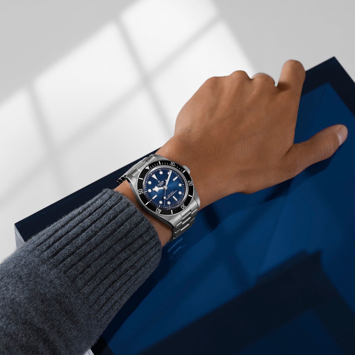 Eine Person, die eine silberne Armbanduhr von Tudor 1926 mit blauem Zifferblatt und blauer Lünette trägt, stützt ihre Hand auf eine glänzende blaue Oberfläche; sie ist mit einem grauen Pullover bekleidet.