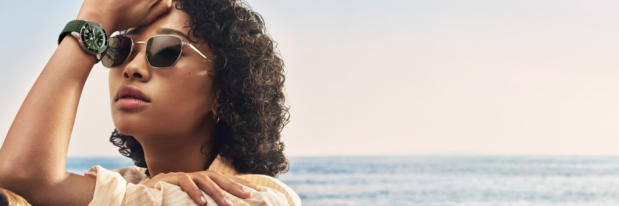Eine Frau mit lockigem Haar, Sonnenbrille und grüner Armbanduhr stützt ihren Arm auf ihren Kopf, während sie an einem sonnigen Tag am Meer steht. Der Himmel ist klar und das Wasser im Hintergrund ist ruhig.