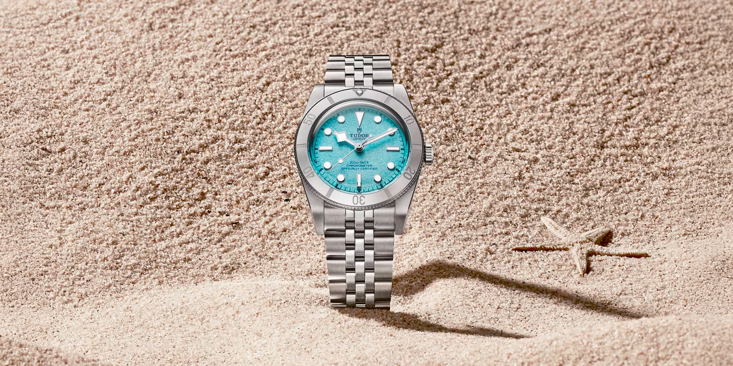 Juwelier-Morawitz-Uhren-Schmuck-Duesseldorf-Blog-TUDOR-Black-Bay-54-Lagoon-Blue-1 Eine TUDOR Black Bay 54 "Lagoon Blue" Armbanduhr mit türkisfarbenem Zifferblatt steht aufrecht im Sand neben einem kleinen Seestern. Ihr silbernes Gehäuse schimmert vor dem strukturierten Sandhintergrund.