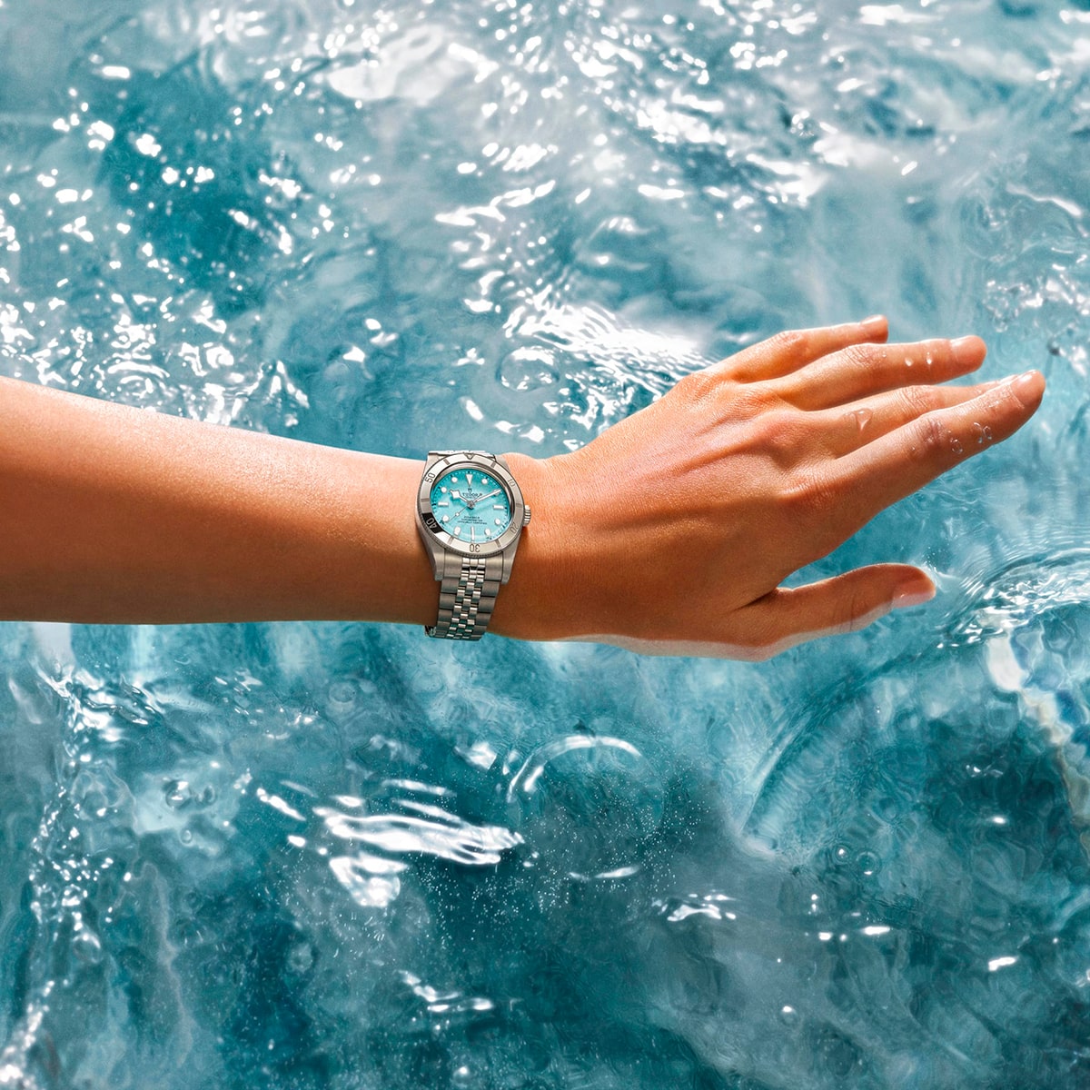Juwelier-Morawitz-Uhren-Schmuck-Duesseldorf-Blog-TUDOR-Black-Bay-54-Lagoon-Blue-2 Eine Hand, die eine silberne TUDOR Black Bay 54 "Lagoon Blue" Armbanduhr mit türkisfarbenem Zifferblatt trägt, schwebt über dem plätschernden blauen Wasser, wobei die Tropfen sowohl an der Haut als auch an der Uhr haften bleiben.