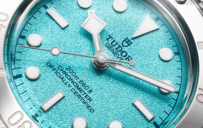 Nahaufnahme einer TUDOR Black Bay 54 "Lagoon Blue" Armbanduhr mit strukturiertem türkisfarbenem Zifferblatt, silbernen Indexen, Datumsfenster bei 3 Uhr und dem Text "Officially Certified". Die Lünette und das Armband aus Edelstahl sind ebenfalls sichtbar.