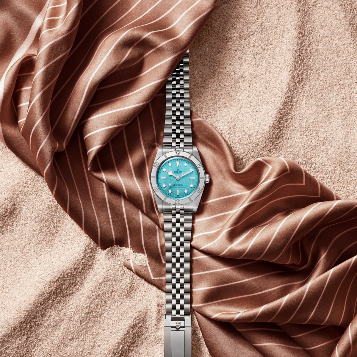 Juwelier-Morawitz-Uhren-Schmuck-Duesseldorf-Blog-TUDOR-Black-Bay-54-Lagoon-Blue-6 Eine TUDOR Black Bay 54 "Lagoon Blue" Armbanduhr mit türkisfarbenem Zifferblatt ruht auf beigem Sand, teilweise umhüllt von glänzendem, braun gestreiftem Stoff. Der Kontrast unterstreicht das elegante Metalldesign vor dem strukturierten und seidigen Hintergrund.