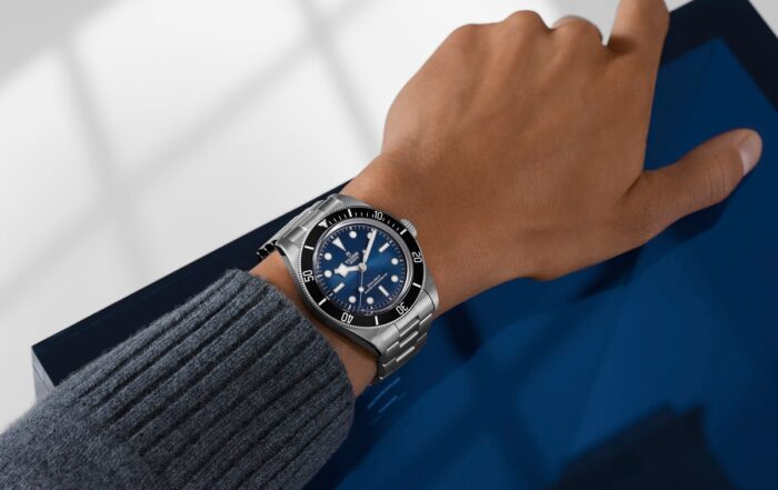 Eine Person, die eine silberne TUDOR Black Bay 68-Edelstahlarmbanduhr mit blauem Zifferblatt und blauer Lünette trägt, stützt ihren Arm auf eine blau glänzende Oberfläche. Die Person ist mit einem grauen gerippten Pullover bekleidet.