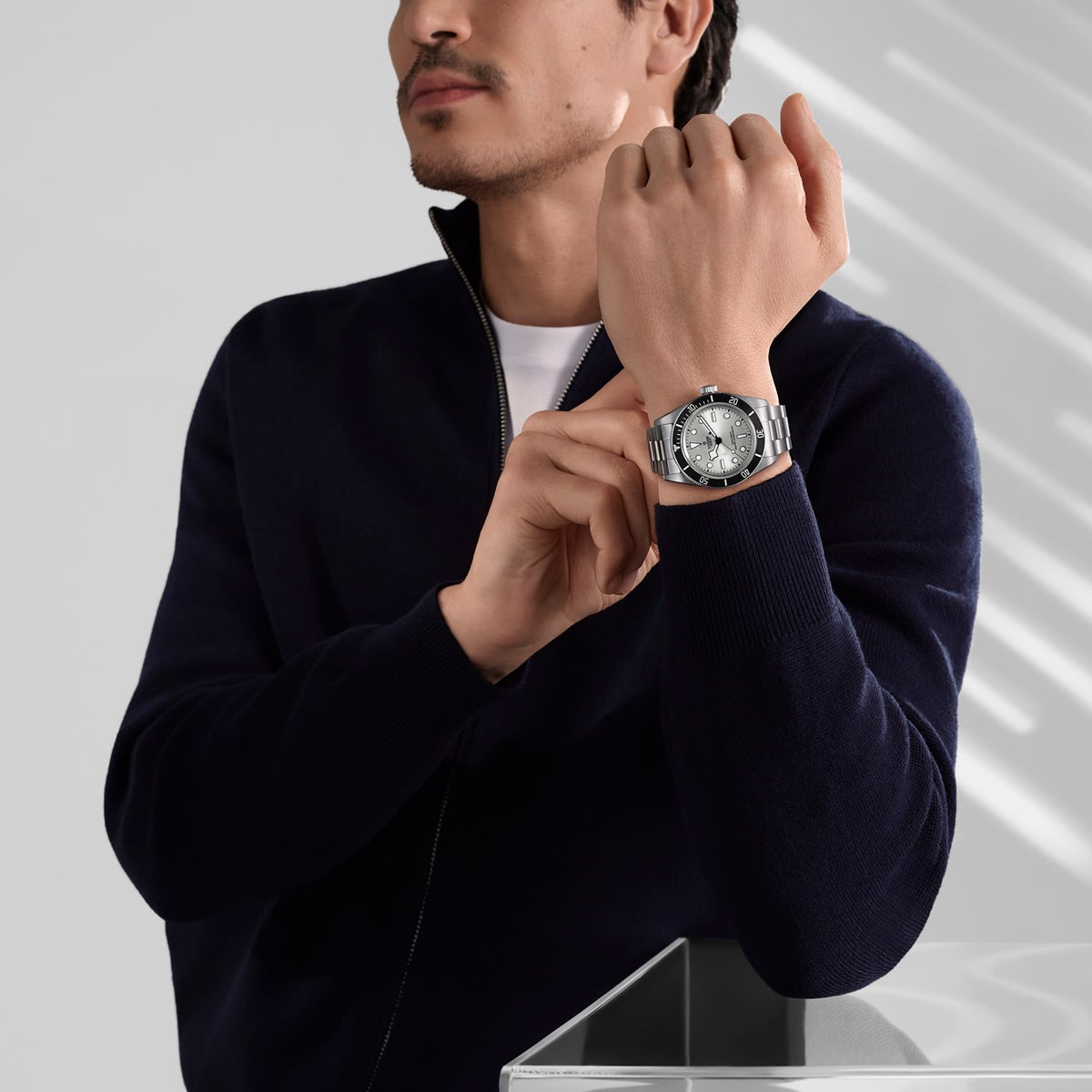 Ein Mann in einem dunklen Pullover mit Reißverschluss rückt eine TUDOR Black Bay 68 an seinem linken Handgelenk zurecht. Er steht neben einer Glasfläche, die wechselnde Muster aus Licht und Schatten auf die Wand hinter ihm wirft. Sein Gesicht ist teilweise sichtbar.