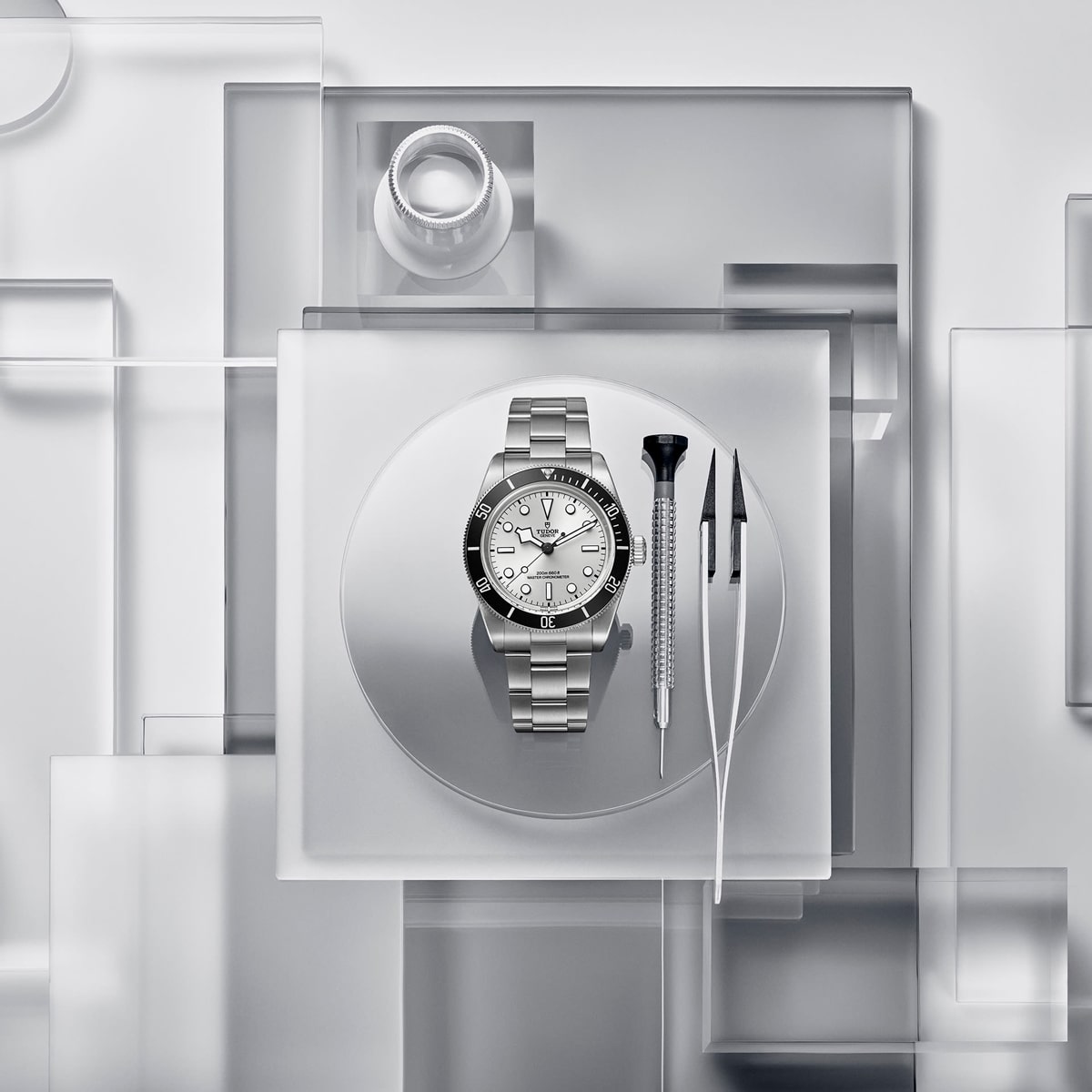 Eine silberne TUDOR Black Bay 68-Armbanduhr wird auf einer Milchglasplatte präsentiert, begleitet von zwei Präzisionswerkzeugen und einer Lupe, die auf transparenten, geometrischen Flächen in minimalistischem Stil angeordnet sind.