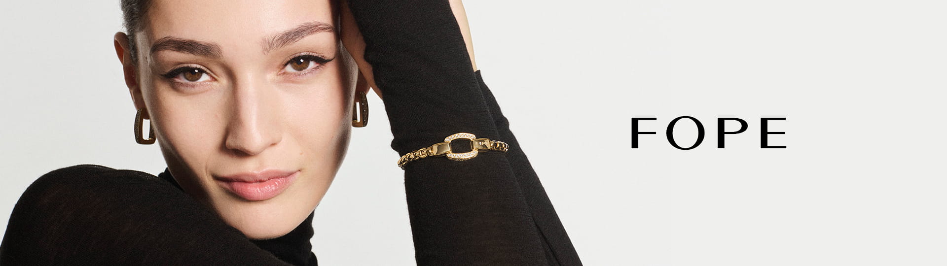 Eine Frau mit braunem Haar, die einen schwarzen Rollkragenpullover, goldene Ohrringe und ein goldenes Fope-Armband trägt, posiert mit erhobenem Arm. Neben ihr erscheint das Wort "FOPE" in schwarzer Schrift auf weißem Hintergrund.